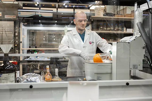A scientist in a white lab coat and safety goggles works with laboratory equipment in a modern chemistry lab. He is operating a rotary evaporator with a round-bottom flask containing an orange substance. Various glassware and chemical containers are visible around him inside a fume hood.